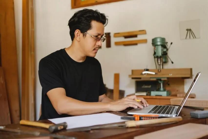 carpenter in workshop, seated, working on laptop